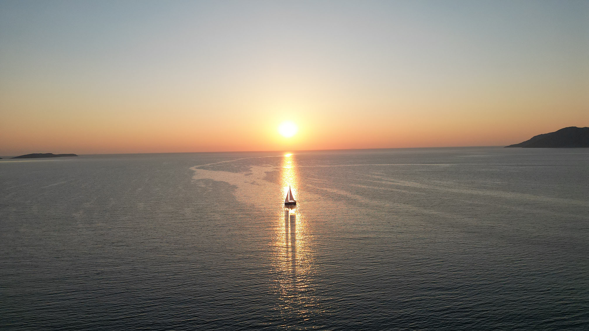 Ein Katamaran gleitet bei Sonnenuntergang auf ruhiger See dem Horizont entgegen. Die goldene Sonne wirft einen schimmernden Lichtstreifen auf die Wasseroberfläche. In der Ferne sind unter einem klaren, von Orange nach Blau wechselnden Himmel schwach kleine Inseln zu erkennen, was dieser Segelreise einen malerischen und ruhigen Charakter verleiht.