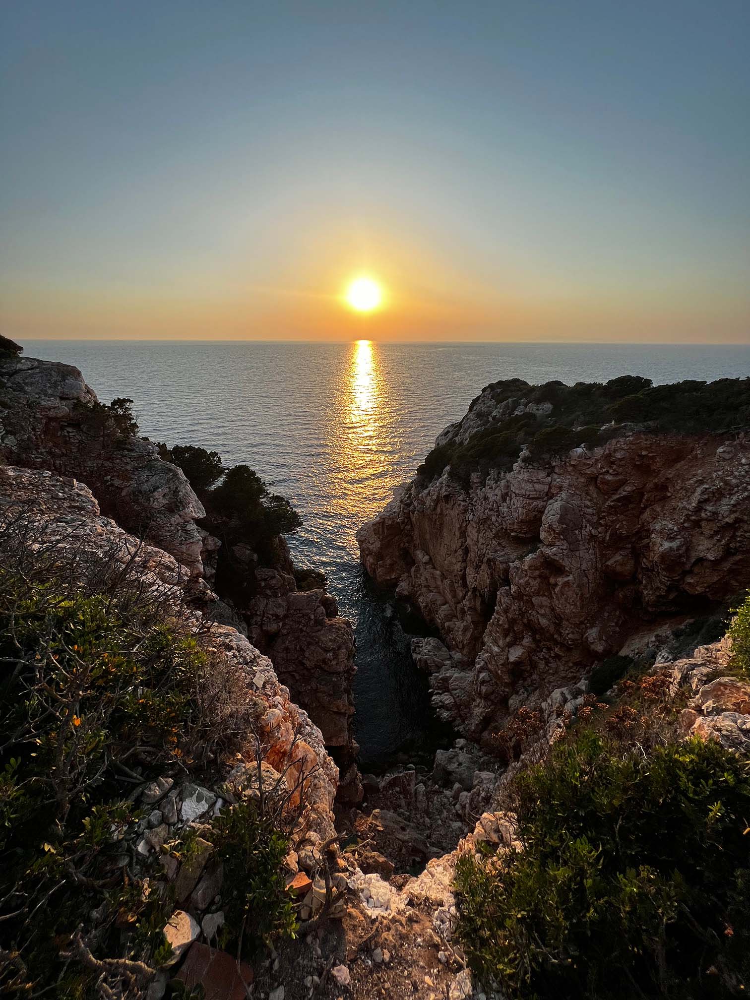 Ein atemberaubender Blick auf einen Sonnenuntergang über dem Meer, gesehen von einer Felsklippe mit grünen Sträuchern. Die Sonne steht tief am Horizont und wirft einen goldenen Widerschein auf das Wasser. Eine Segelyacht segelt sanft entlang der schroffen Klippenränder und umrahmt eine schmale Bucht, die auf das offene Meer hinausführt.