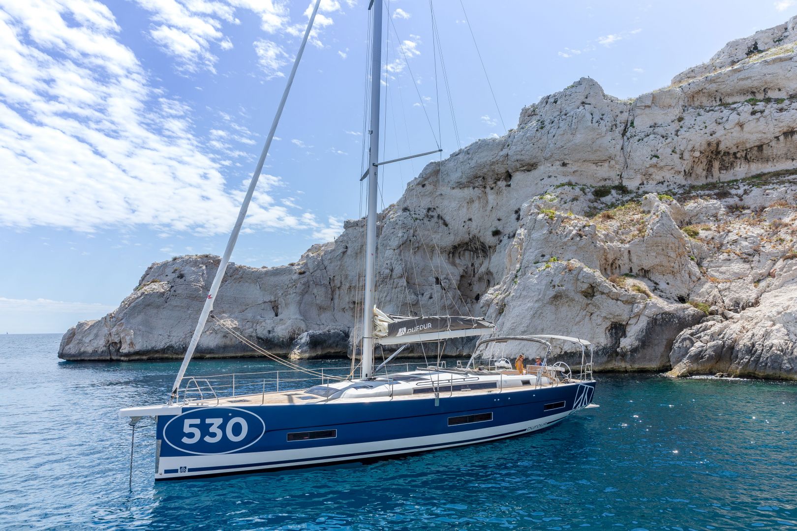 Ein blaues Segelboot mit der Nummer 530 auf der Seite liegt in der Nähe einer felsigen, weißen Klippe vor Anker. Das klare, blaue Wasser umgibt das Boot und darüber ist ein teilweise bewölkter Himmel zu sehen. Auf dem Boot sind ein paar Leute, die ihr Yachtcharter-Erlebnis genießen.