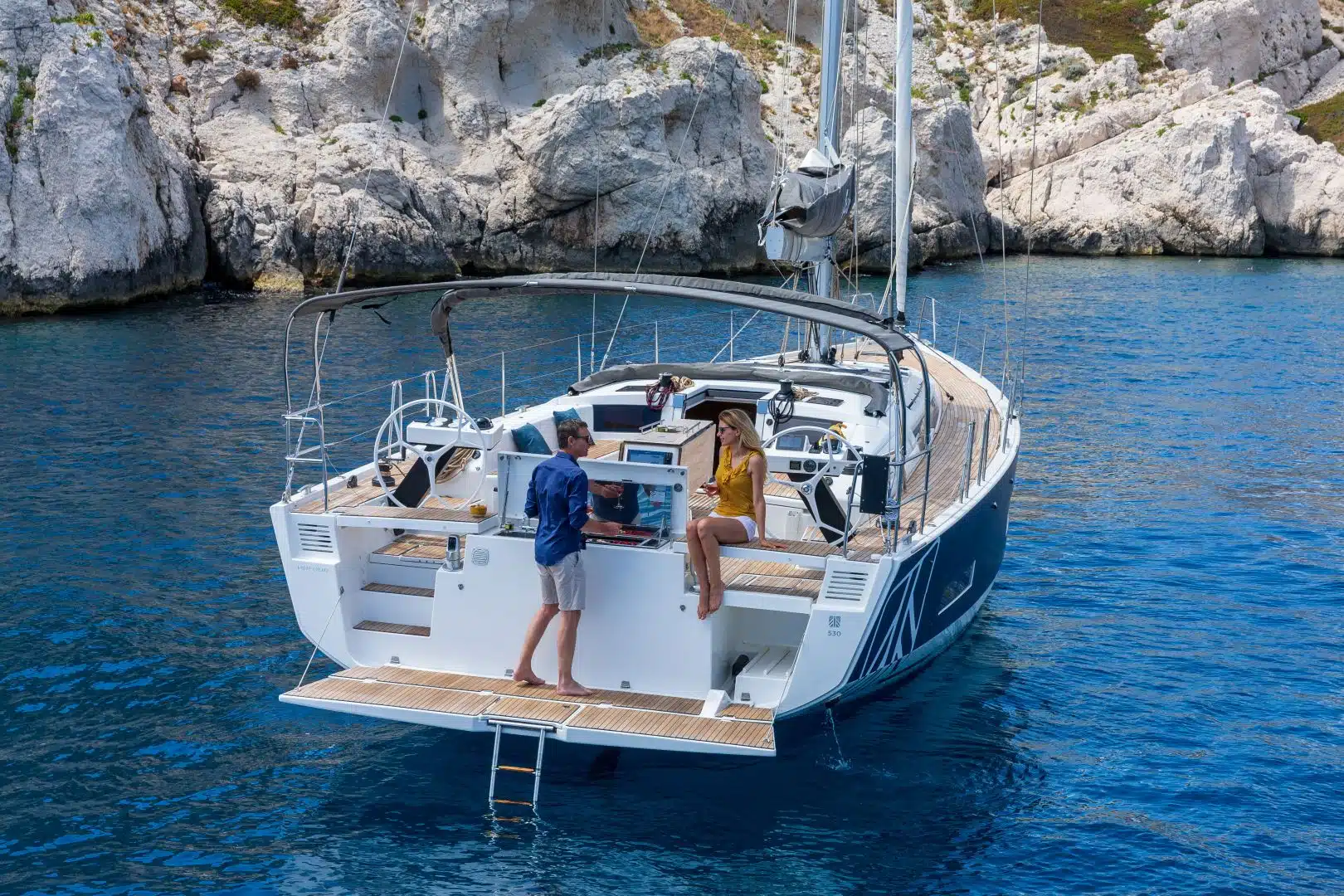 Ein Mann und eine Frau sind auf einem Segelboot in klarem, blauem Wasser, mit felsigen Klippen im Hintergrund. Die Frau sitzt am Rand, während der Mann am Steuer steht. Das elegante Yachtcharter-Schiff ist mit modernen Annehmlichkeiten ausgestattet, perfekt für ein unvergessliches Seeabenteuer.
