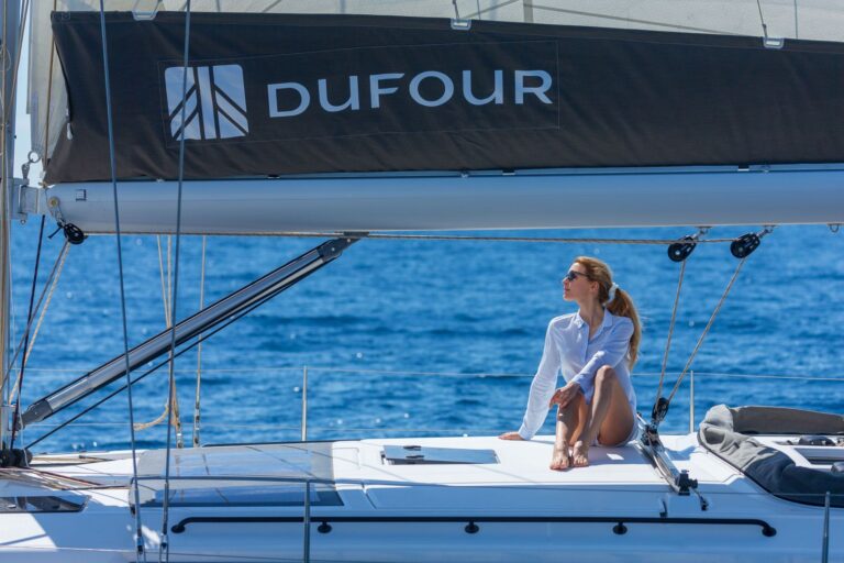 Eine Frau in weißem Hemd und Sonnenbrille sitzt auf dem Deck einer Yacht und genießt das sonnige Wetter. Das Segelboot „DUFOUR“ gleitet über das ruhige blaue Wasser. Sie wirkt entspannt, genießt die Sonne und verkörpert die unbeschwerte Eleganz von Yachtcharter-Abenteuern.