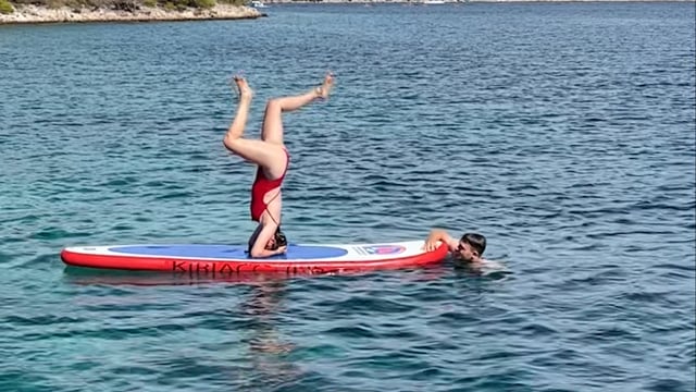Eine Person im roten Badeanzug macht einen Kopfstand auf einem Paddleboard in ruhigem, klarem Wasser. Eine andere Person mit Schwimmbrille schwimmt in der Nähe und stützt das Paddleboard teilweise. Im Hintergrund ist eine entfernte Küstenlinie unter einem sonnigen Himmel zu sehen.
