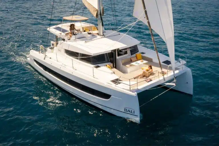 Ein weißer Youngline-Katamaran mit der Aufschrift „BALI“ auf den Seitensegeln auf klarem, blauem Wasser. Drei Personen entspannen auf dem Boot; eine auf dem Oberdeck und zwei auf dem Hauptdeck. Das Boot, perfekt für einen Segeltörn, hat Sonnenliegen und einen offenen Sitzbereich vorne.