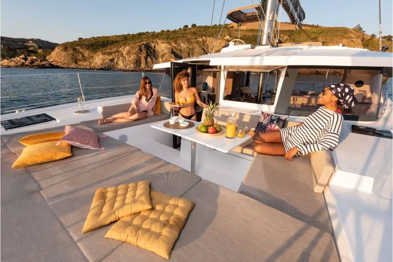 Vier Personen entspannen auf einem luxuriösen Youngline-Katamaran mit Bergen und Wasser im Hintergrund. Das Deck ist mit gepolsterten Sitzgelegenheiten und einem Tisch mit Obst und Getränken ausgestattet. Die Atmosphäre ist sonnig und heiter und lässt auf einen gemütlichen Ausflug auf dem Wasser schließen.
