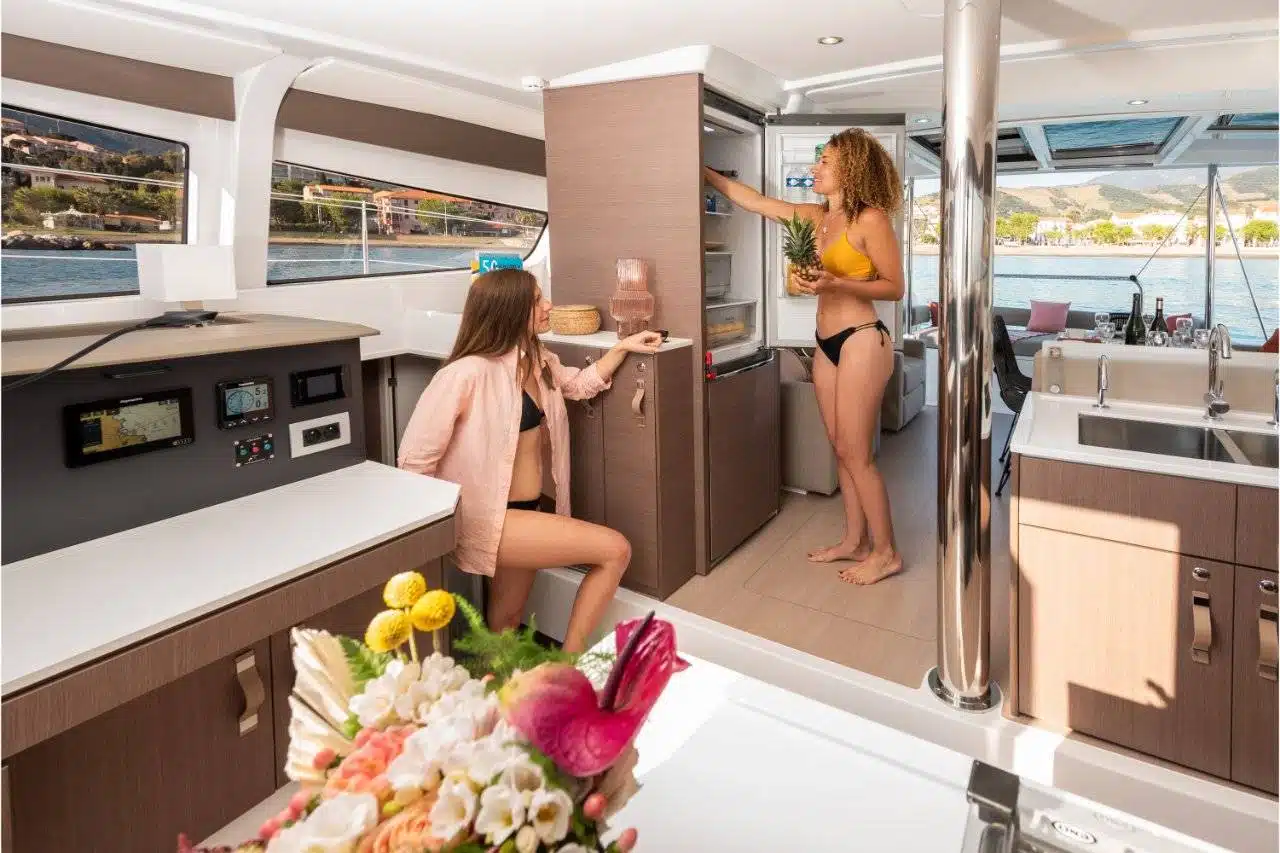 Zwei Frauen in Badeanzügen stehen in der Küche eines modernen Youngline Katamarans. Eine steht und legt eine Ananas in einen Schrank, während die andere sitzt und nach einem Snack greift. Es gibt Bedienfelder, Blumen auf der Theke und durch die Fenster blickt man auf das Meer und die Berge.
