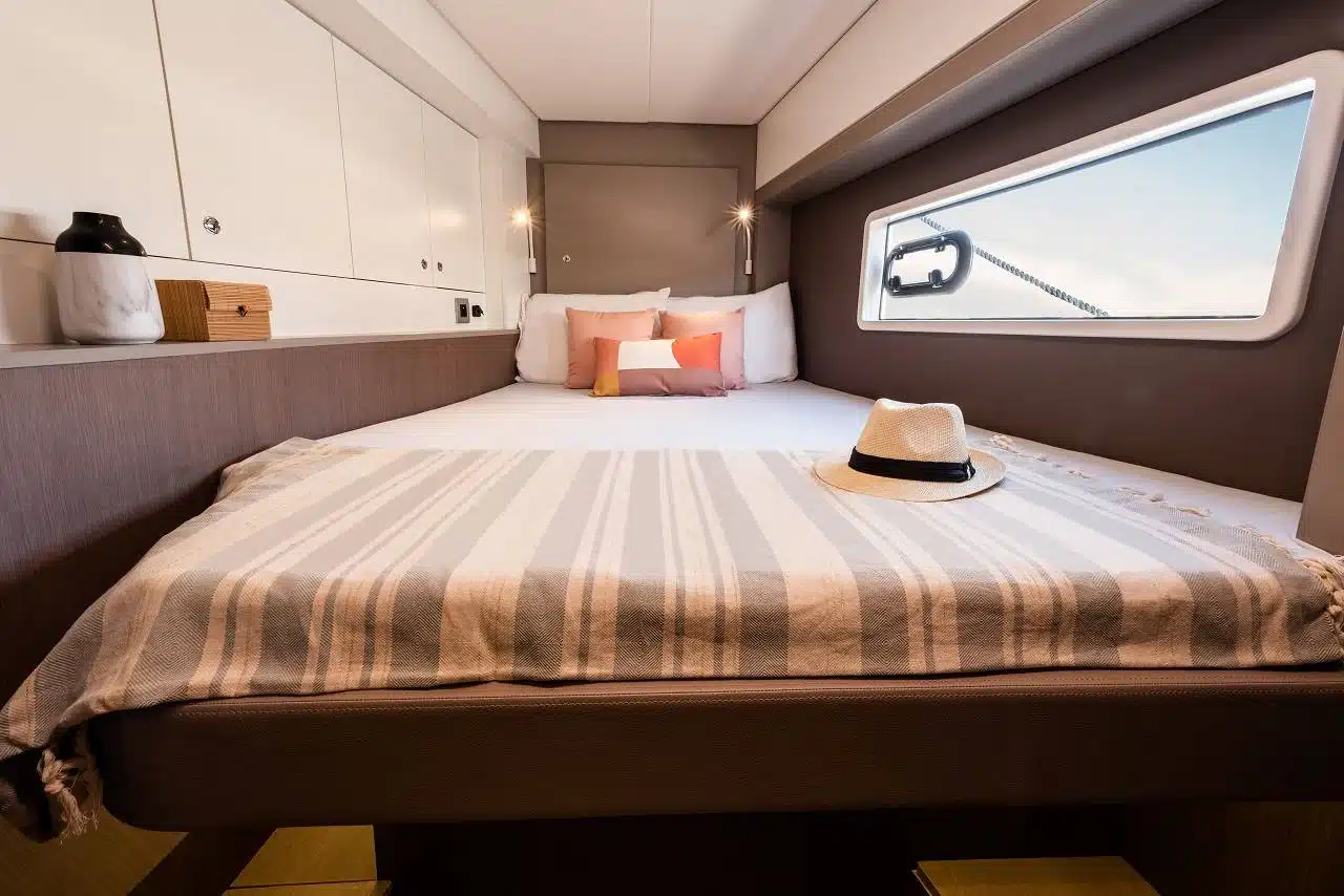 Ein ordentlich gemachtes Bett mit gestreifter Bettwäsche in einer Kabine mit Holz- und Weißakzenten. Auf dem Bett liegen ein Hut und ein buntes Kissen. Die Kabine verfügt über Einbauschränke, ein Fenster mit Blick auf den Youngline Katamaran Segeltörn und gezielte Beleuchtung am Kopfende des Bettes.