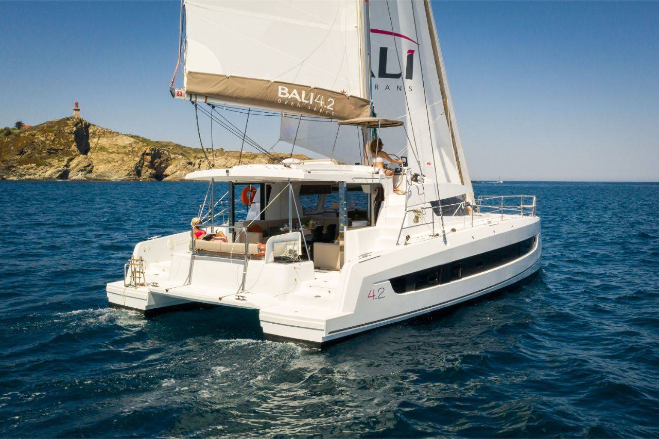 Ein weißer Katamaran namens „Bali 4.2“ segelt auf klarem, blauem Wasser in der Nähe einer felsigen Küste mit einem Leuchtturm. Das Boot hat Segel gesetzt und mehrere Personen sind an Deck zu sehen, die den sonnigen Tag genießen. Wahrscheinlich besteigen sie einen Youngline Katamaran Segeltörn.