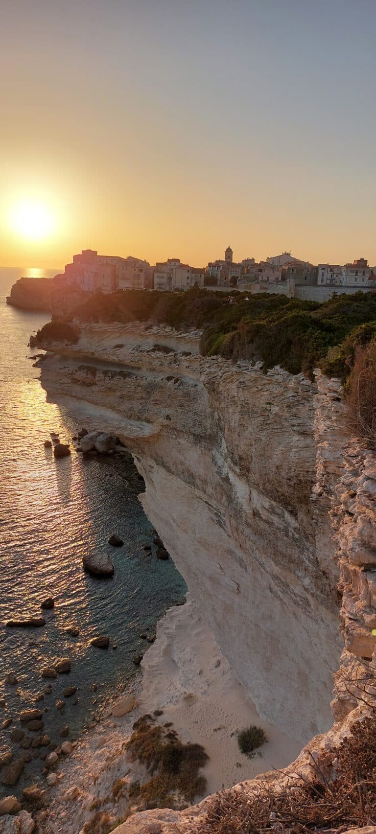 Ein malerischer Küstenblick bei Sonnenuntergang zeigt eine schroffe beige Klippe, die zu einem Sandstrand und klarem, ruhigem Wasser mit verstreuten Felsen führt. Eine Ansammlung von Gebäuden steht auf den Klippen und hebt sich während eines Segelurlaubs auf Sardinien gegen die goldenen Farbtöne der untergehenden Sonne ab.