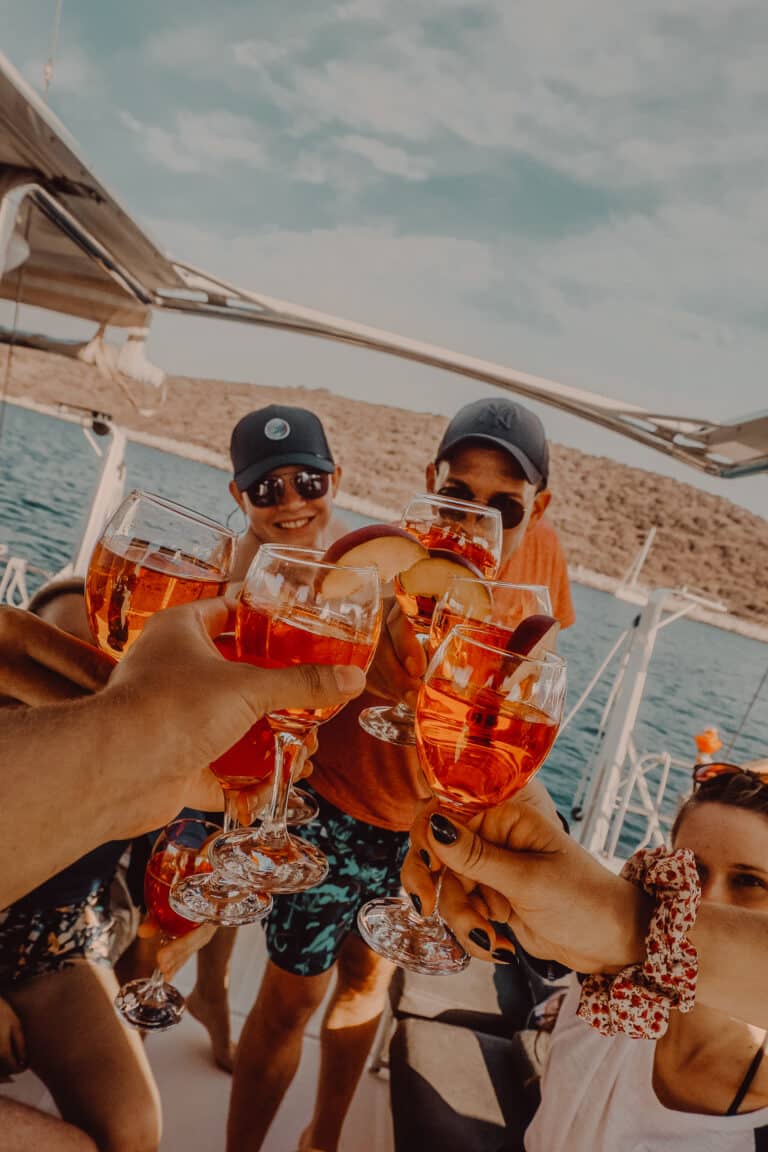 Eine Gruppe von Menschen auf einem Boot stößt mit einem leuchtend orangefarbenen Getränk an und genießt ihren Segelurlaub. Sie lächeln und genießen das sonnige Wetter. Im Hintergrund sind das Meer und eine hügelige Küste zu sehen.