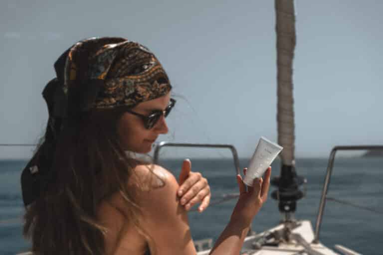 Eine Frau mit buntem Kopftuch und Sonnenbrille sitzt während ihrer Segelreise in Griechenland auf einem Boot und cremt ihre Schulter mit Sonnencreme ein. In der anderen Hand hält sie eine Tube Sonnencreme. Im Hintergrund ist das Meer zu sehen.