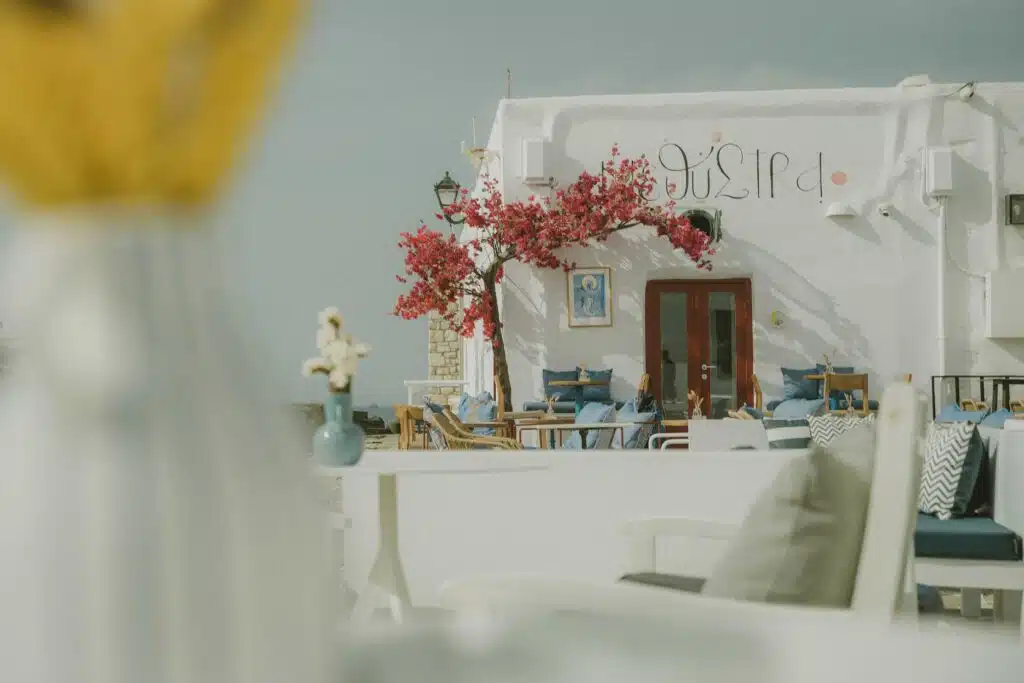 Ein charmantes Café im Freien verfügt über weiße Tische und Stühle mit blauen Kissen. Im Vordergrund steht ein leuchtender Bougainvillea-Baum mit rosa Blüten vor einem weißen Gebäude mit einem griechischen Schild. Die Szene strahlt eine heitere, mediterrane Atmosphäre aus, die an ein Abenteuer von Segelreise Griechenland erinnert.