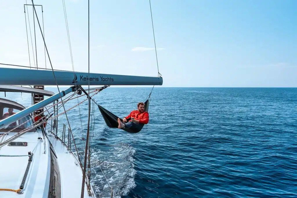 Eine Person in roter Jacke und schwarzen Shorts entspannt sich in einer Hängematte, die am Baum einer Segelyacht mit dem Aufdruck „Kekeris Yachts“ hängt. Das Boot segelt auf einem ruhigen blauen Meer unter klarem Himmel und genießt den ultimativen Segeltörn.
