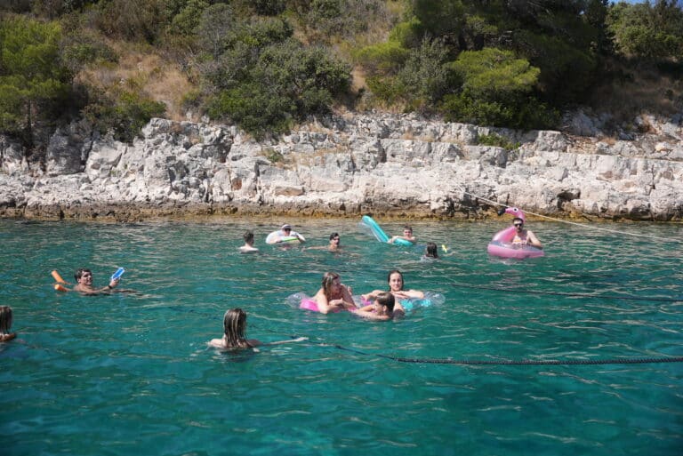 Eine Gruppe von Menschen genießt einen sonnigen Tag im klaren blauen Wasser nahe einer felsigen Küste. Einige schwimmen, während andere bunte Schwimmflügel und Poolnudeln benutzen. Bäume und Sträucher sind auf dem felsigen Hintergrund zu sehen, was an Mitsegeln in Kroatien erinnert. Die Szene ist lebhaft und entspannt.