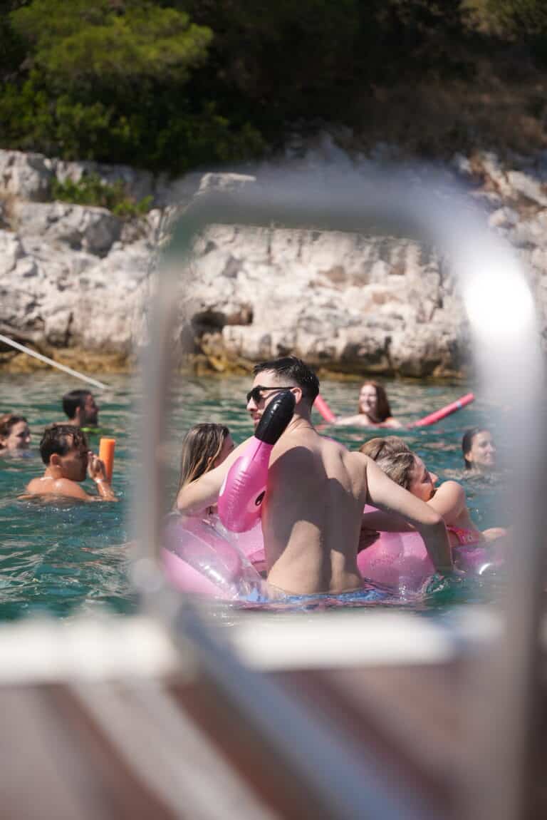 Eine Gruppe von Menschen genießt einen sonnigen Tag im Wasser, im Hintergrund felsige Klippen. Einige treiben auf aufblasbaren Spielzeugen, darunter einem großen rosafarbenen. Das Bild, das an ein Abenteuer von Mitsegeln Kroatien erinnert, wird im Vordergrund von einem unscharfen Geländer eingerahmt.