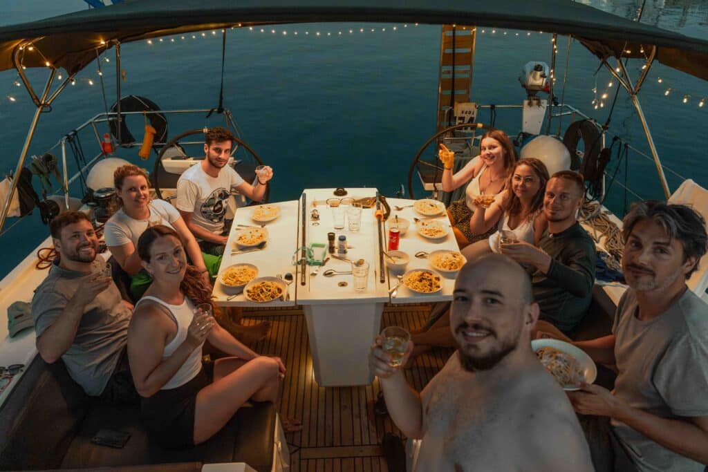 Eine Gruppe von zehn Personen sitzt auf einer Segeltour um einen Tisch und genießt gemeinsam eine Mahlzeit. Auf dem Tisch stehen Teller mit Pasta und Getränken. Die Gruppe scheint sich zu unterhalten und zu lächeln, während festliche Lichterketten eine warme Atmosphäre schaffen, während die Sonne über dem Wasser untergeht. Mitsegeln in Kleingruppen: Gemeinsam die Meere erkunden