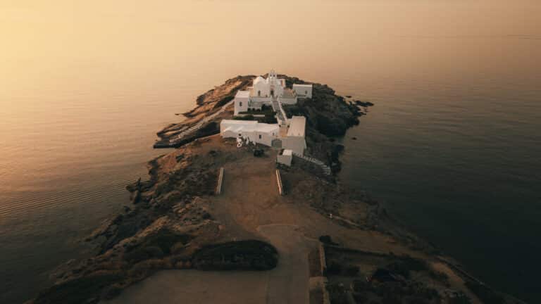 Luftaufnahme einer Halbinsel bei Sonnenuntergang. Weiße Gebäude drängen sich an der Spitze der Landform, umgeben von ruhigem, reflektierendem Wasser. Die Szene ist in ein warmes, goldenes Licht getaucht. Spärliche Vegetation und felsiges Gelände führen zu den Gebäuden – ein idyllischer Anblick für diejenigen auf einer Segeltour.