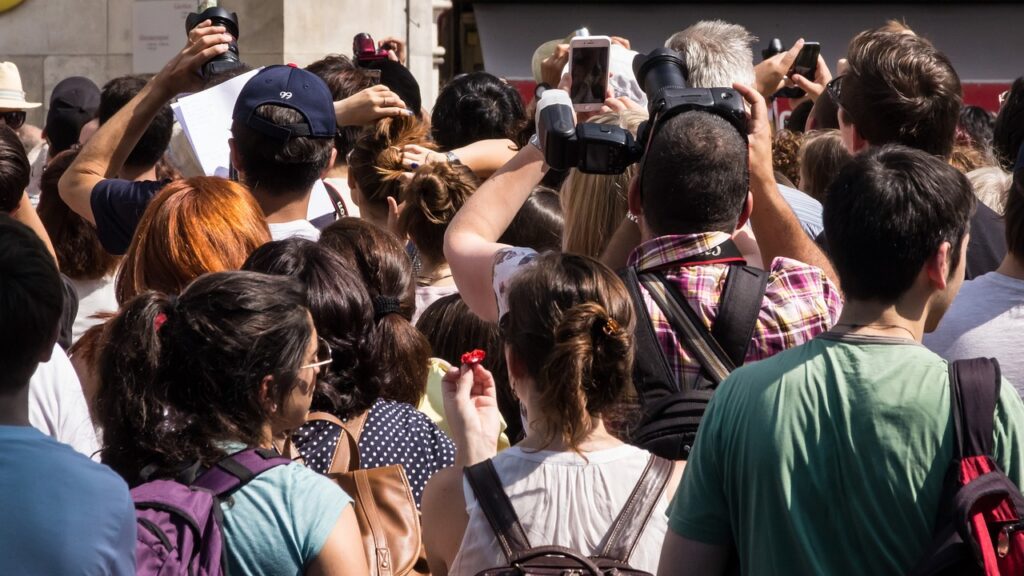 Eine Menschenmenge, von hinten und oben gesehen, viele halten Kameras und Smartphones hoch und fokussieren auf etwas außerhalb des Sichtfelds. Die Szene lässt auf eine geschäftige öffentliche Veranstaltung oder eine Touristenattraktion schließen, die von Übertourismus geplagt ist. Die Personen sind unterschiedlichen Alters und leger gekleidet. Das ist Overtourism.