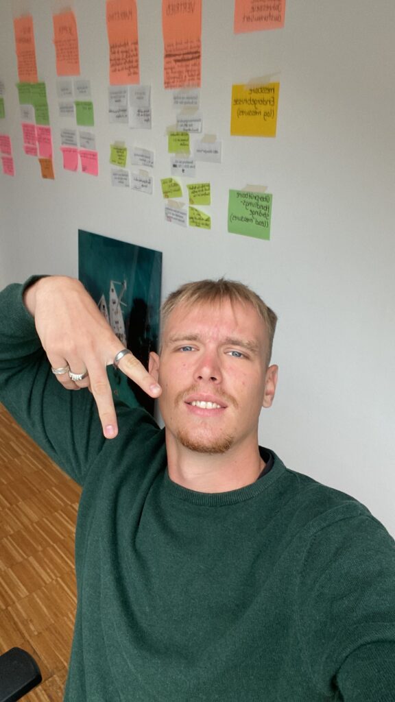 Eine Person mit kurzem hellem Haar und hellem Bart trägt einen grünen Pullover und macht mit der Hand ein Peace-Zeichen. Sie befindet sich in einem Gebäude vor einer Wand voller bunter Haftnotizen und Papiere, vermutlich von Teamprojekten. Der Boden ist aus Holz und die Person lächelt leicht.