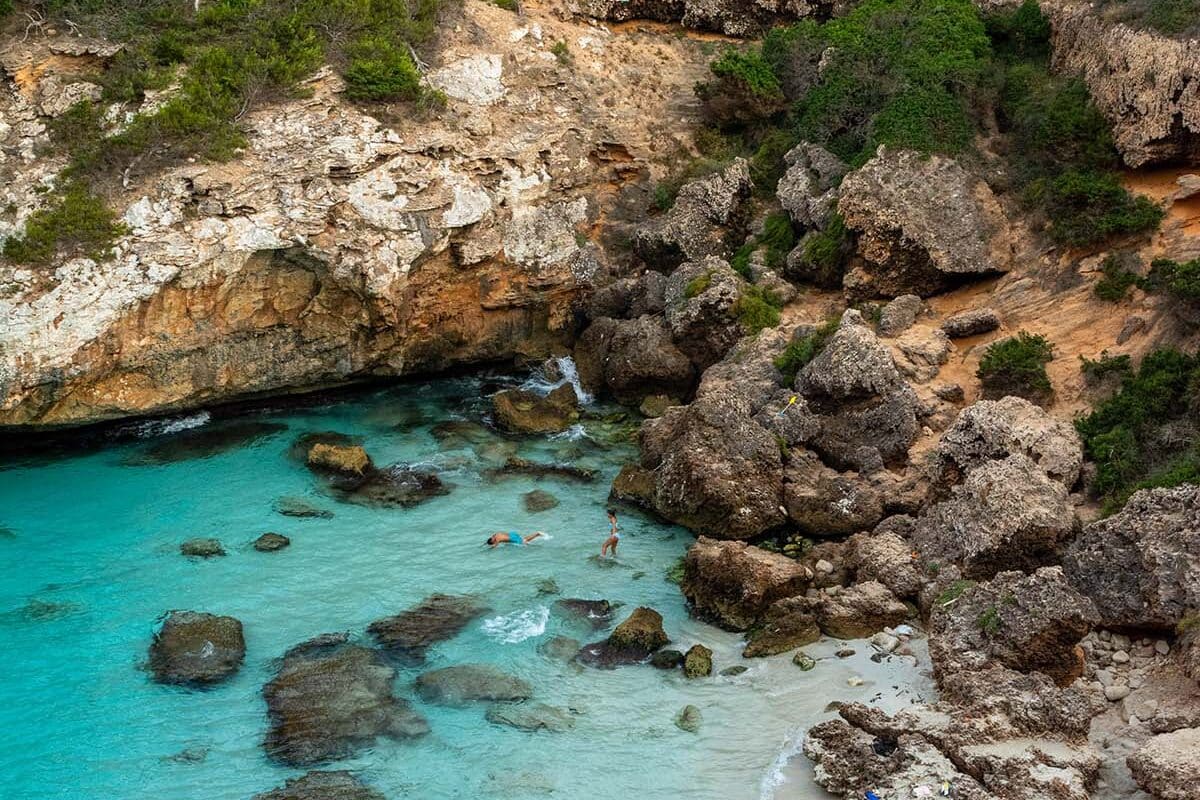 Eine versteckte Bucht mit türkisfarbenem Wasser in Mallorca, umgeben von schroffen Klippen und viel Grün, wo die Leute schwimmen und den abgeschiedenen Strandbereich von Segeltörn genießen.
