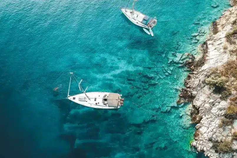 Luftaufnahme von zwei weißen Booten, die in der Nähe der felsigen Küste eines kristallklaren blauen Meeres vor Anker liegen, perfekt für einen Segeltörn.
