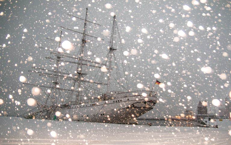 Sailing ship docked in snowy Stralsund harbor, Germany, during winter snowfall.